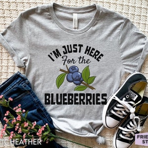 Op de afbeelding: Een grijs T-shirt met de tekst "I'm just here for the blueberries" en een afbeelding van blauwe bessen aan een tak.
