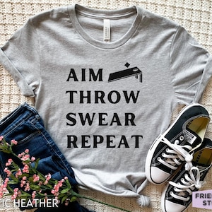 Könnte beinhalten: Ein graues T-Shirt mit schwarzem Text, der "AIM THROW SWEAR REPEAT" lautet, und einer schwarzen Silhouette eines Cornhole-Bretts. Das Shirt liegt auf einer weißen und grauen strukturierten Oberfläche.