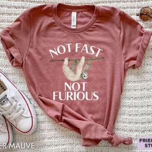 May include: A heather mauve short-sleeve t-shirt featuring a graphic of a sloth hanging from a branch with the white text "NOT FAST NOT FURIOUS" printed above and below the sloth. The shirt has a crew neckline and a casual, relaxed fit.
