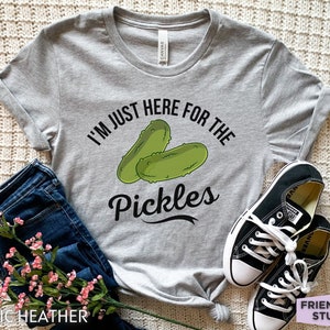 Puede incluir: Camiseta gris jaspeado con el texto "I'M JUST HERE FOR THE PICKLES" y un gráfico de dos pepinillos verdes. La camiseta se combina con vaqueros azules y zapatillas negras.