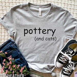 Puede incluir: Una camiseta gris con las palabras "pottery (and cats)" en negro. La camiseta se combina con vaqueros azules, zapatillas deportivas blancas y negras y una ramita de flores rosas. La camiseta es de material athletic heather.