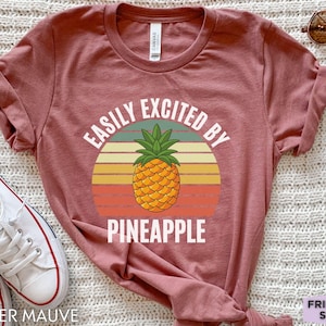 May include: Heather mauve t-shirt with a retro design featuring a pineapple and the text "EASILY EXCITED BY PINEAPPLE". The shirt has rolled-up sleeves and is paired with white sneakers and sunglasses. The words "Friend Tee Studio" are also visible.