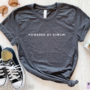 Pode incluir: Uma camiseta cinza heather escura com o texto "POWERED BY KIMCHI" impresso em branco na frente. A camiseta está colocada sobre uma superfície de madeira branca com um par de tênis pretos, óculos de sol e uma planta suculenta em vaso.