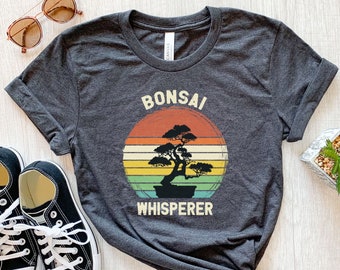 May include: A dark gray heather t-shirt featuring a graphic of a bonsai tree centered within a multi-colored sunset circle. The text BONSAI is printed in white above the tree, and the word WHISPERER is printed in white below the tree graphic.