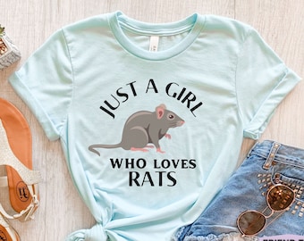 May include: A light blue short-sleeve t-shirt featuring a graphic of a grey rat centered between the black text that reads JUST A GIRL WHO LOVES RATS. The shirt has a crew neckline and is displayed with casual accessories including sandals and denim.
