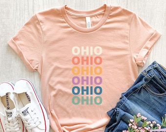 Könnte beinhalten: Ein kurzärmeliges T-Shirt in Pfirsich-Melange mit dem Wort OHIO, das sechsmal vertikal untereinander gedruckt ist, wobei jedes Wort eine andere Farbe hat, darunter Creme, Orange, Gelb, Lila, Blau und Grün, kombiniert mit weißen Turnschuhen und blauen Jeans.