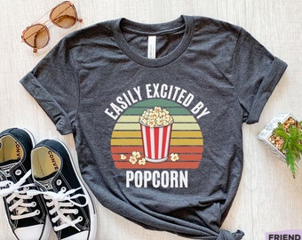 Może przedstawiać: Ciemnoszary melanżowy t-shirt z grafiką przedstawiającą czerwono-biały kubełek na popcorn na tle zachodzącego słońca w stylu retro. Nad grafiką znajduje się napis "EASILY EXCITED BY", a poniżej wydrukowane jest słowo "POPCORN".