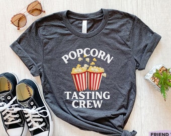 Może przedstawiać: Ciemnoszary melanżowy t-shirt z okrągłym dekoltem, ozdobiony grafiką przedstawiającą dwa czerwono-białe kubełki na popcorn oraz białym napisem POPCORN TASTING CREW wykonanym pogrubioną czcionką bezszeryfową nad i pod kubełkami.