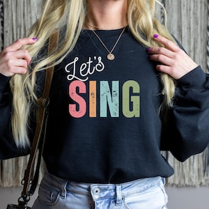 Op de afbeelding: Een zwart sweatshirt met lange mouwen met de witte cursieve tekst Let's boven het woord SING in blokletters met een verweerd, meerkleurig lettertype. De letters van SING zijn gekleurd in roze, oranje, lichtblauw en saliegroen.
