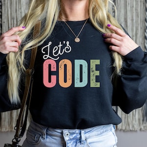 Op de afbeelding: Zwart sweatshirt met de tekst "Let's CODE" in witte letters en kleurrijke blokletters. Het woord "CODE" is roze, oranje, blauw en groen. Het sweatshirt heeft een casual stijl.