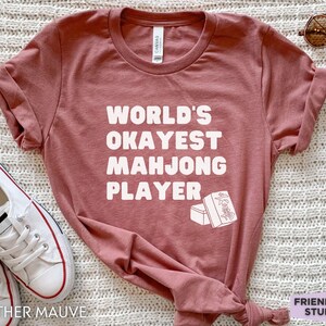 May include: A heather mauve t-shirt with the text "WORLD'S OKAYEST MAH JONG PLAYER" in white. The shirt features a white illustration of two mahjong tiles.