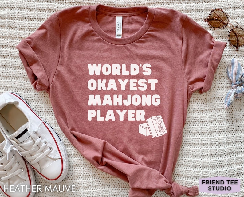 May include: A heather mauve t-shirt with the text "WORLD'S OKAYEST MAH JONG PLAYER" in white. The shirt features a white illustration of two mahjong tiles.