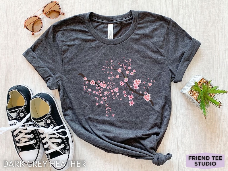 May include: Dark grey heather t-shirt with a pink cherry blossom branch design. The t-shirt is shown on a wooden surface with a pair of black sneakers and a small potted plant.