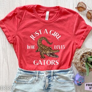 Puede incluir: Camiseta rojo jaspeado con mangas enrolladas, con el texto "JUST A GIRL WHO LOVES GATORS" y una ilustraci&oacute;n detallada de un caim&aacute;n. La camiseta se combina con pantalones cortos de mezclilla y gafas de sol.