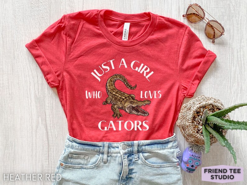 Puede incluir: Camiseta rojo jaspeado con mangas enrolladas, con el texto "JUST A GIRL WHO LOVES GATORS" y una ilustraci&oacute;n detallada de un caim&aacute;n. La camiseta se combina con pantalones cortos de mezclilla y gafas de sol.