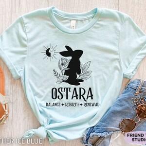 Puede incluir: Una camiseta azul claro con un gr&aacute;fico negro de un conejo rodeado de flores y un s&iacute;mbolo de sol y luna. Debajo del gr&aacute;fico, el texto dice OSTARA, seguido de las palabras BALANCE, REBIRTH y RENEWAL separadas por s&iacute;mbolos de diamantes.