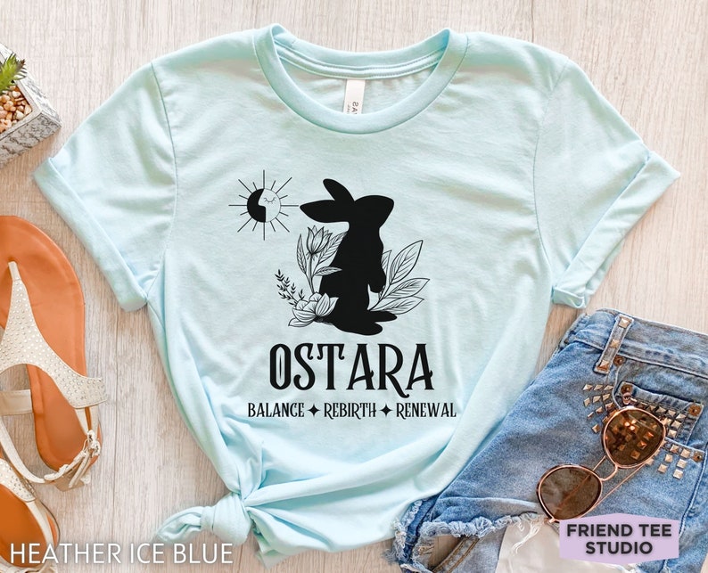 Puede incluir: Una camiseta azul claro con un gr&aacute;fico negro de un conejo rodeado de flores y un s&iacute;mbolo de sol y luna. Debajo del gr&aacute;fico, el texto dice OSTARA, seguido de las palabras BALANCE, REBIRTH y RENEWAL separadas por s&iacute;mbolos de diamantes.