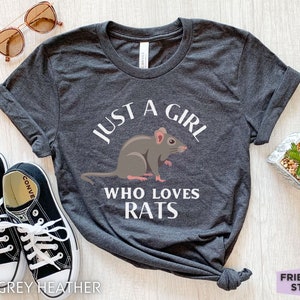 May include: A dark grey heather t-shirt featuring a graphic of a grey rat and the white text "JUST A GIRL WHO LOVES RATS" printed on the front. The shirt has short sleeves and a crew neckline, displayed with black and white canvas sneakers.