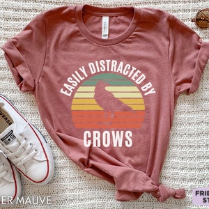 May include: A heather mauve t-shirt with a vintage-style graphic of a crow in front of a sunset. The text "Easily Distracted By Crows" is printed on the shirt.