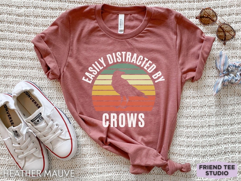 May include: A heather mauve t-shirt with a vintage-style graphic of a crow in front of a sunset. The text "Easily Distracted By Crows" is printed on the shirt.