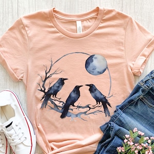 May include: A peach-colored t-shirt featuring a watercolor design of three black crows perched on a branch, with a full moon in the background. The design is enclosed in a gray circle. The shirt is paired with blue jeans and white sneakers.