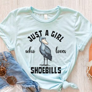 Puede incluir: Una camiseta azul claro con el texto "Just a girl who loves shoebills" en negro. Una ilustraci&oacute;n de dibujos animados de un ave zancuda se encuentra en el centro de la camiseta.