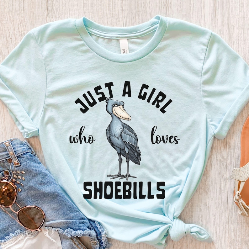 Puede incluir: Una camiseta azul claro con el texto "Just a girl who loves shoebills" en negro. Una ilustraci&oacute;n de dibujos animados de un ave zancuda se encuentra en el centro de la camiseta.