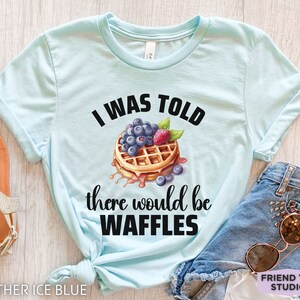 May include: Heather ice blue t-shirt with the text "I WAS TOLD there would be WAFFLES" and a waffle graphic topped with blueberries and a raspberry. The shirt is paired with denim shorts and sunglasses.