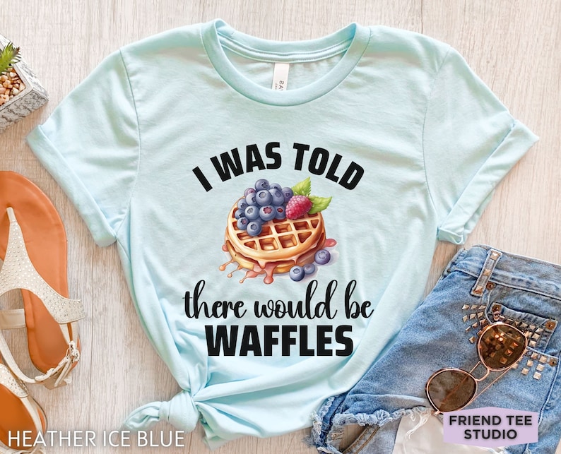 May include: Heather ice blue t-shirt with the text "I WAS TOLD there would be WAFFLES" and a waffle graphic topped with blueberries and a raspberry. The shirt is paired with denim shorts and sunglasses.
