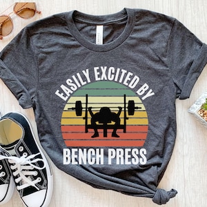 Peut inclure: T-shirt gris foncé avec l'inscription blanche "EASILY EXCITED BY BENCH PRESS". Le graphisme représente une silhouette d'haltérophile soulevant des poids sur un fond de coucher de soleil rayé jaune, vert, orange et rouge.