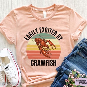 Op de afbeelding: Een perzikkleurig T-shirt met een retro-grafische afbeelding van een rivierkreeft en de tekst "Easily Excited By Crawfish".