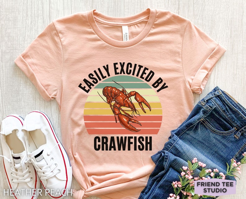 Op de afbeelding: Een perzikkleurig T-shirt met een retro-grafische afbeelding van een rivierkreeft en de tekst "Easily Excited By Crawfish".