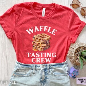 May include: Heather red t-shirt with the words "WAFFLE TASTING CREW" in white. A stack of waffles with syrup is in the center. The shirt is paired with denim shorts. Sunglasses and a small plant are in the background.