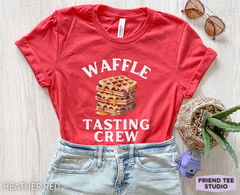 May include: Heather red t-shirt with the words "WAFFLE TASTING CREW" in white. A stack of waffles with syrup is in the center. The shirt is paired with denim shorts. Sunglasses and a small plant are in the background.