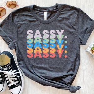 Puede incluir: Camiseta gris oscuro con la palabra "SASSY" en un diseño apilado y colorido. La palabra está en una variedad de colores, incluyendo morado, verde, azul, naranja y rojo. La camiseta es de estilo casual, adecuada para el uso diario.
