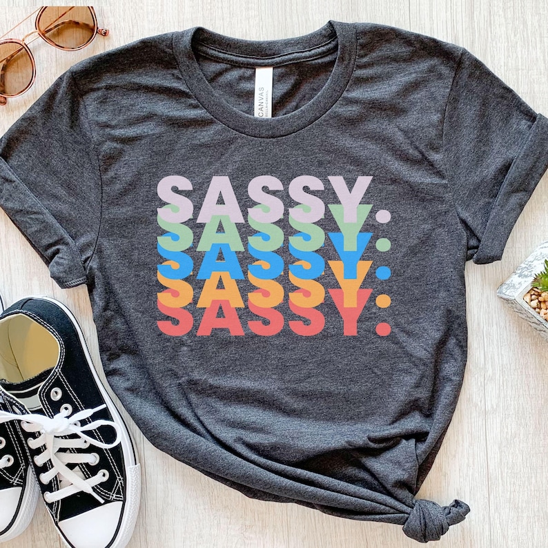 May include: Dark gray t-shirt with the word "SASSY" in a stacked, colorful design. The word is in a variety of colors, including purple, green, blue, orange, and red. The shirt is a casual style, suitable for everyday wear.