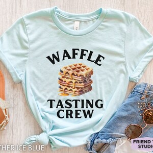 May include: Heather ice blue t-shirt with the words "WAFFLE TASTING CREW" in black above and below a stack of waffles with syrup and blueberries. The shirt is styled with denim shorts and sunglasses.