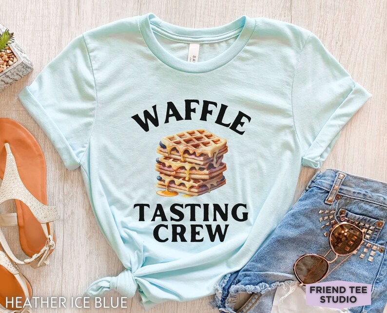 May include: Heather ice blue t-shirt with the words "WAFFLE TASTING CREW" in black above and below a stack of waffles with syrup and blueberries. The shirt is styled with denim shorts and sunglasses.