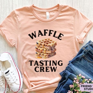 May include: A peach-colored t-shirt with rolled-up sleeves featuring the text "WAFFLE TASTING CREW" above and below a stack of waffles with syrup and blueberries. White sneakers and blue jeans are also in the image.