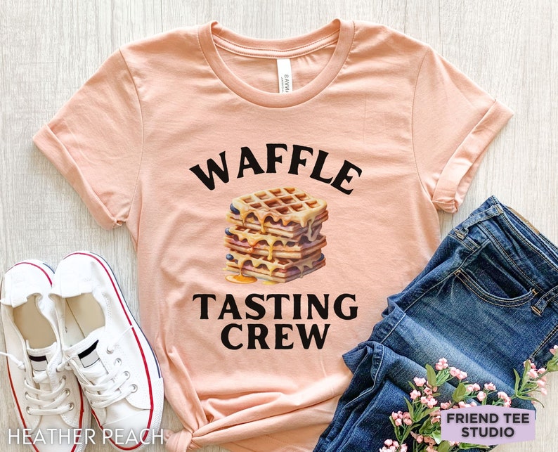 May include: A peach-colored t-shirt with rolled-up sleeves featuring the text "WAFFLE TASTING CREW" above and below a stack of waffles with syrup and blueberries. White sneakers and blue jeans are also in the image.