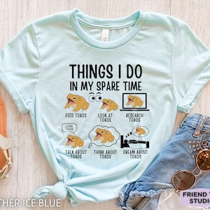 May include: Light blue t-shirt with the text "THINGS I DO IN MY SPARE TIME" and illustrations of toads. The shirt also features the phrases "FEED TOADS", "LOOK AT TOADS", "RESEARCH TOADS", "TALK ABOUT TOADS", "THINK ABOUT TOADS", and "DREAM ABOUT TOADS".