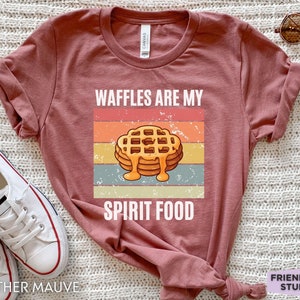 Pu&ograve; includere: T-shirt color malva con grafica di waffle con sciroppo e la scritta "WAFFLES ARE MY SPIRIT FOOD". Il design &egrave; ambientato su uno sfondo a righe in stile retr&ograve;.