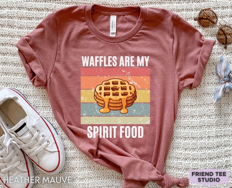 Pu&ograve; includere: T-shirt color malva con grafica di waffle con sciroppo e la scritta "WAFFLES ARE MY SPIRIT FOOD". Il design &egrave; ambientato su uno sfondo a righe in stile retr&ograve;.