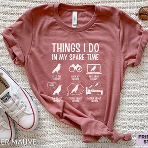 K&ouml;nnte beinhalten: Ein T-Shirt in Heide-Mauve mit einer wei&szlig;en Grafik, auf der "Things I do in my spare time" steht und Aktivit&auml;ten im Zusammenhang mit Kr&auml;hen aufgelistet sind.