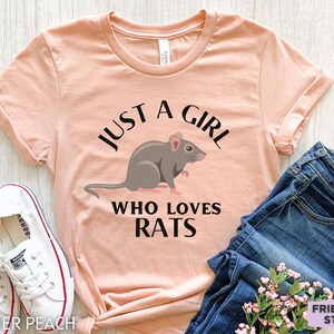 May include: A heather peach short-sleeve t-shirt featuring a graphic of a grey rat and the black text "JUST A GIRL WHO LOVES RATS" printed on the front. The shirt is displayed alongside white canvas sneakers and blue denim jeans.