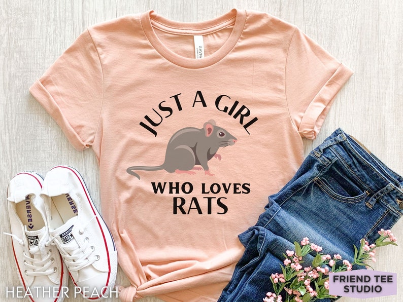 May include: A heather peach short-sleeve t-shirt featuring a graphic of a grey rat and the black text "JUST A GIRL WHO LOVES RATS" printed on the front. The shirt is displayed alongside white canvas sneakers and blue denim jeans.