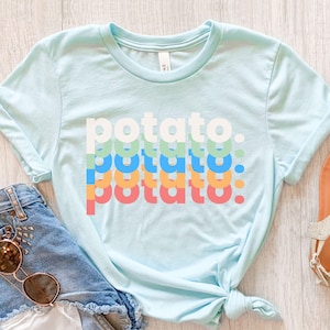 May include: A light blue short-sleeve t-shirt featuring the word potato printed in four stacked rows of different colors including white, light green, blue, orange, and pink. The text is lowercase and includes a period at the end of each line.