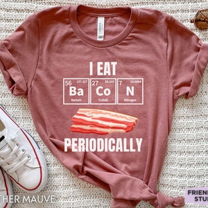 Puede incluir: Camiseta color malva con la frase "I EAT BACON PERIODICALLY" en blanco. El dise&ntilde;o incluye una tira de tocino y los elementos de la tabla peri&oacute;dica Ba, Co y N. Prenda casual.