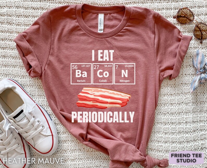 Puede incluir: Camiseta color malva con la frase "I EAT BACON PERIODICALLY" en blanco. El dise&ntilde;o incluye una tira de tocino y los elementos de la tabla peri&oacute;dica Ba, Co y N. Prenda casual.