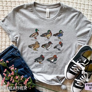 Peut inclure: Un t-shirt gris chin&eacute; avec une impression graphique de huit esp&egrave;ces de canards diff&eacute;rentes, accompagn&eacute;es de leurs noms respectifs : Mallard, Wood, Baikal Teal, Gadwall, Eurasian Teal, Redhead, Rosy Billed Pochard et Red Crested Pochard.